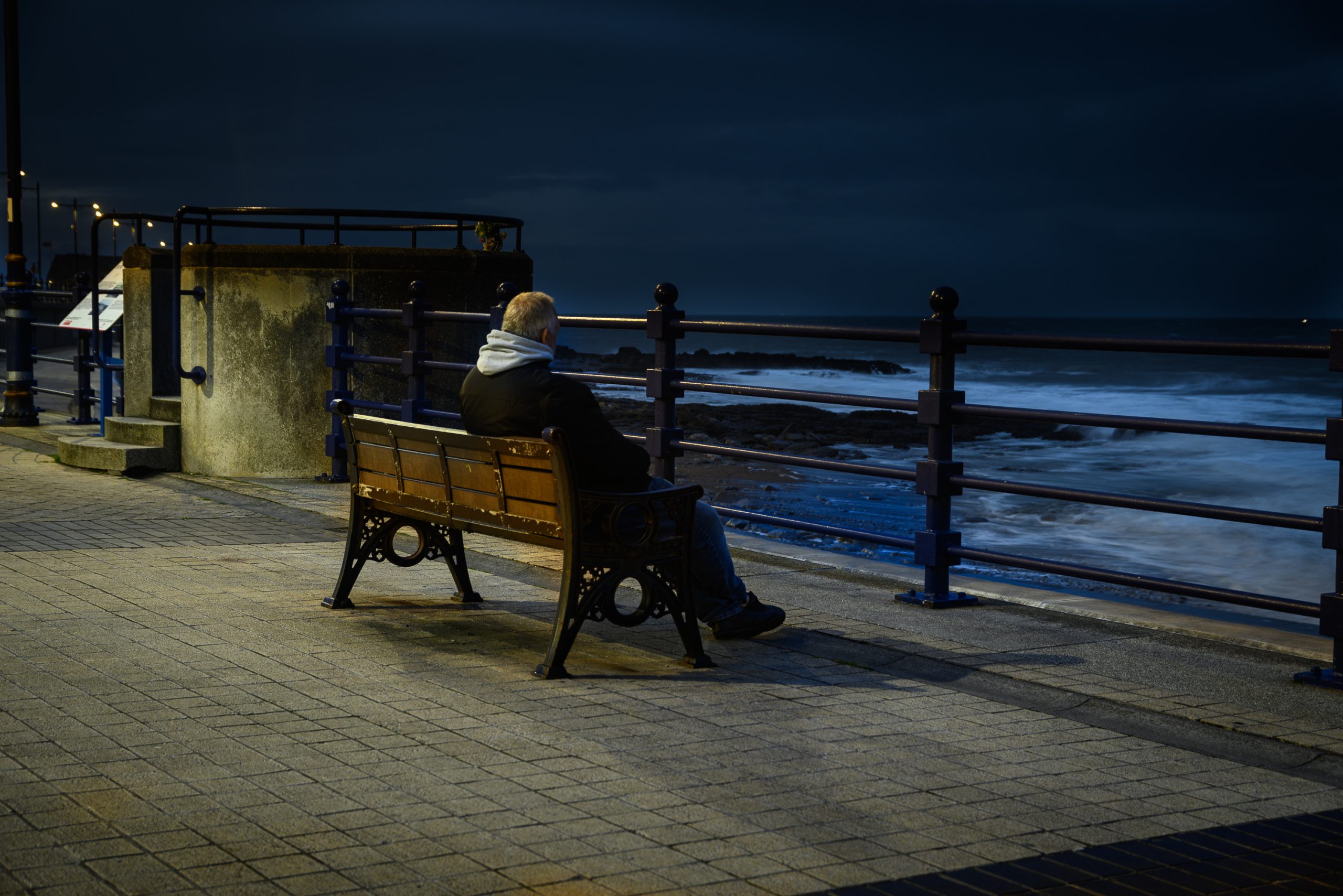 A person in a dark jacket and gray hood sits on a wooden bench along a waterfront promenade at night, gazing toward the ocean with a metal railing nearby.