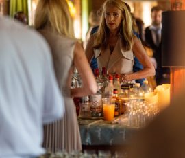 Blonde bartender in a beige sleeveless top behind a bottle-filled bar, preparing drinks in a warmly lit room. Blonde bartender in a beige sleeveless top behind a bottle-filled bar, preparing drinks in a warmly lit room.