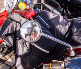 Close-up of a red motorcycle with protective gear draped over the seat; round mirror shows a person in reflection. Close-up of a red motorcycle with protective gear draped over the seat; round mirror shows a person in reflection