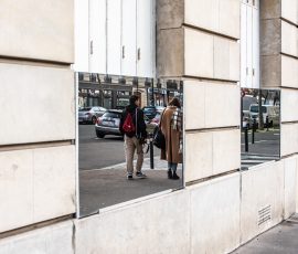 People walking past a mirror, a man wearing a black jacket and light trousers while carrying a camera, and a woman wearing a long light coat with a scarf over her shoulder. People walking past a mirror, a man wearing a black jacket and light trousers while carrying a camera, and a woman wearing a long light coat with a scarf over her shoulder.