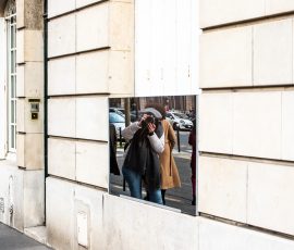 Person photographing themselves in a mirror/window reflection on a stone building, wearing a gray coat and black scarf, urban street scene Person photographing themselves in a mirror/window reflection on a stone building, wearing a gray coat and black scarf, urban street scene